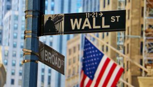 Straßenschilder für Wall Street und Broad Street an einer Kreuzung, mit einer verschwommenen amerikanischen Flagge und hohen Gebäuden im Hintergrund - eine Anspielung auf den Einfluss von Bitcoin auf das traditionelle Finanzwesen.