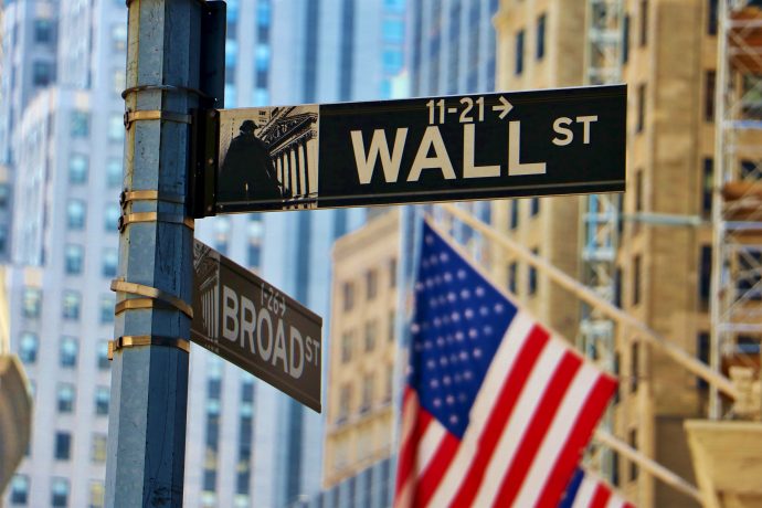 Straßenschilder für Wall Street und Broad Street an einer Kreuzung, mit einer verschwommenen amerikanischen Flagge und hohen Gebäuden im Hintergrund - eine Anspielung auf den Einfluss von Bitcoin auf das traditionelle Finanzwesen.