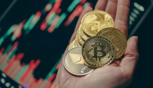 Eine Hand hält mehrere physische Krypto-Münzen, darunter Bitcoin, Ethereum und andere, mit einem digitalen Aktienchart im verschwommenen Hintergrund.