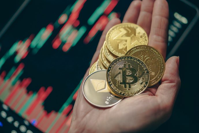 Eine Hand hält mehrere physische Krypto-Münzen, darunter Bitcoin, Ethereum und andere, mit einem digitalen Aktienchart im verschwommenen Hintergrund.