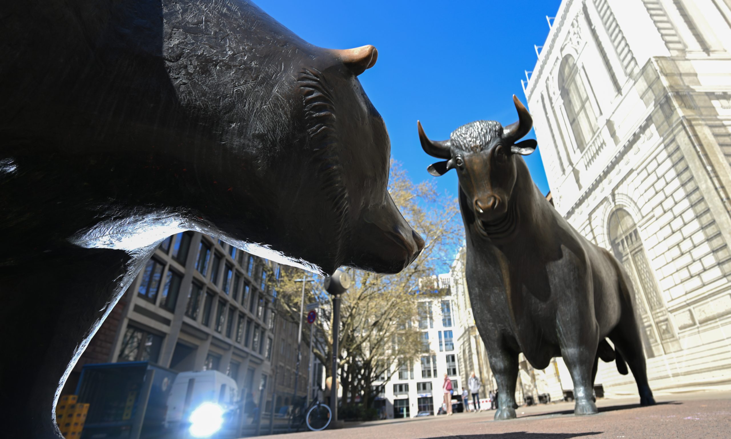 Eine Bären- und eine Bullenstatue aus Bronze stehen sich auf einer Straße in der Stadt gegenüber und symbolisieren gegensätzliche Trends auf dem Aktienmarkt und den Aufstieg der Blockchain-Technologie. Gebäude und ein klarer Himmel sorgen für eine moderne Kulisse.