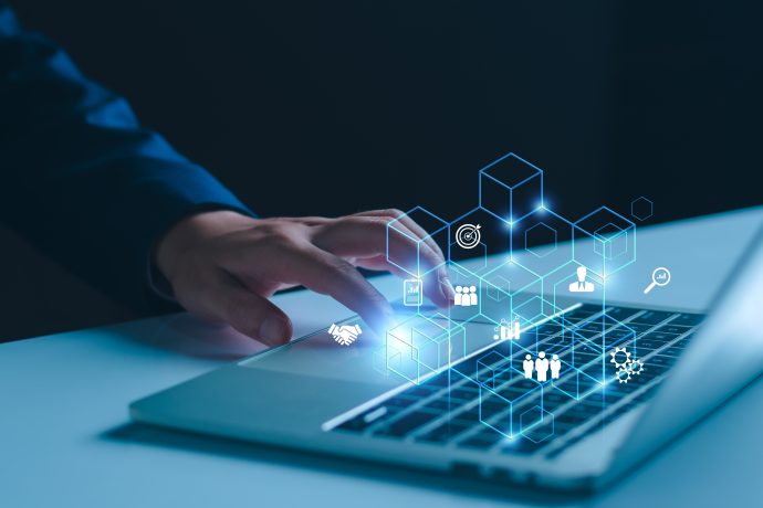 Eine Person tippt auf einer Laptoptastatur, während über dem Gerät digitale Geschäfts- und Technologiesymbole wie Würfel und Menschen sowie Blockchain-Symbole projiziert werden.