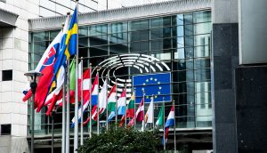 Vor einem modernen Glasgebäude mit dem Emblem des Europäischen Parlaments an der Fassade sind mehrere europäische Nationalflaggen und die EU-Flagge zu sehen, die ein visuelles Zeichen für die Einheit und Zusammenarbeit in Europa setzen.
