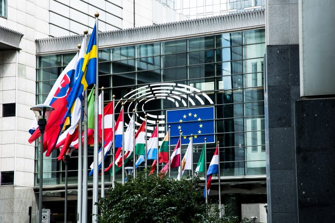Vor einem modernen Glasgebäude mit dem Emblem des Europäischen Parlaments an der Fassade sind mehrere europäische Nationalflaggen und die EU-Flagge zu sehen, die ein visuelles Zeichen für die Einheit und Zusammenarbeit in Europa setzen.