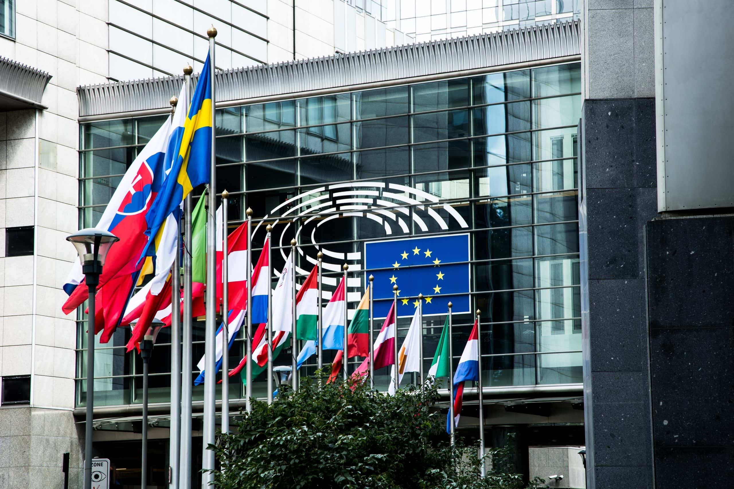Vor einem modernen Glasgebäude mit dem Emblem des Europäischen Parlaments an der Fassade sind mehrere europäische Nationalflaggen und die EU-Flagge zu sehen, die ein visuelles Zeichen für die Einheit und Zusammenarbeit in Europa setzen.