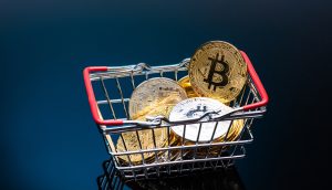 Ein kleiner Einkaufskorb, gefüllt mit physischen Darstellungen von Kryptowährungsmünzen, einschließlich Bitcoin, steht auf einer dunklen, reflektierenden Oberfläche.