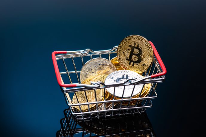 Ein kleiner Einkaufskorb, gefüllt mit physischen Darstellungen von Kryptowährungsmünzen, einschließlich Bitcoin, steht auf einer dunklen, reflektierenden Oberfläche.