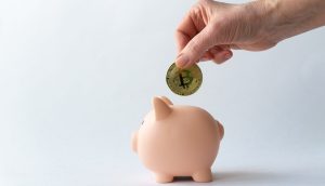 Eine Hand legt eine Bitcoin-Münze in ein rosafarbenes Sparschwein vor einem schlichten weißen Hintergrund, das modernes Sparen symbolisiert - sei es für die Zukunft oder als Teil der Planung neben traditionellen Renten wie der Witwenrente.