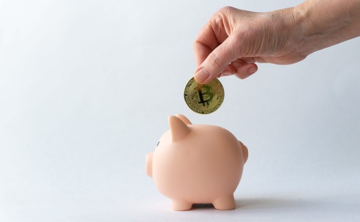 Eine Hand legt eine Bitcoin-Münze in ein rosafarbenes Sparschwein vor einem schlichten weißen Hintergrund, das modernes Sparen symbolisiert - sei es für die Zukunft oder als Teil der Planung neben traditionellen Renten wie der Witwenrente.