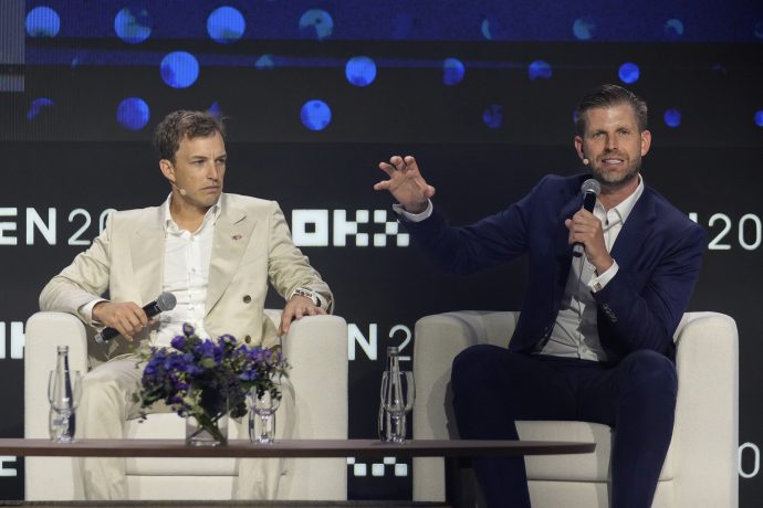 Zach Witkoff und Eric Trump in Anzügen sitzen während einer WLFI-Podiumsdiskussion auf der Bühne; einer spricht in ein Mikrofon, während der andere zuhört. Ein Tisch mit Blumen und Wasserflaschen steht vor ihnen.