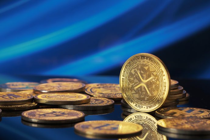 Mehrere goldfarbene XRP-Münzen der Kryptowährung Ripple sind auf einer reflektierenden Oberfläche verstreut, wobei ein blauer abstrakter Hintergrund die Bedeutung von XRP hervorhebt.