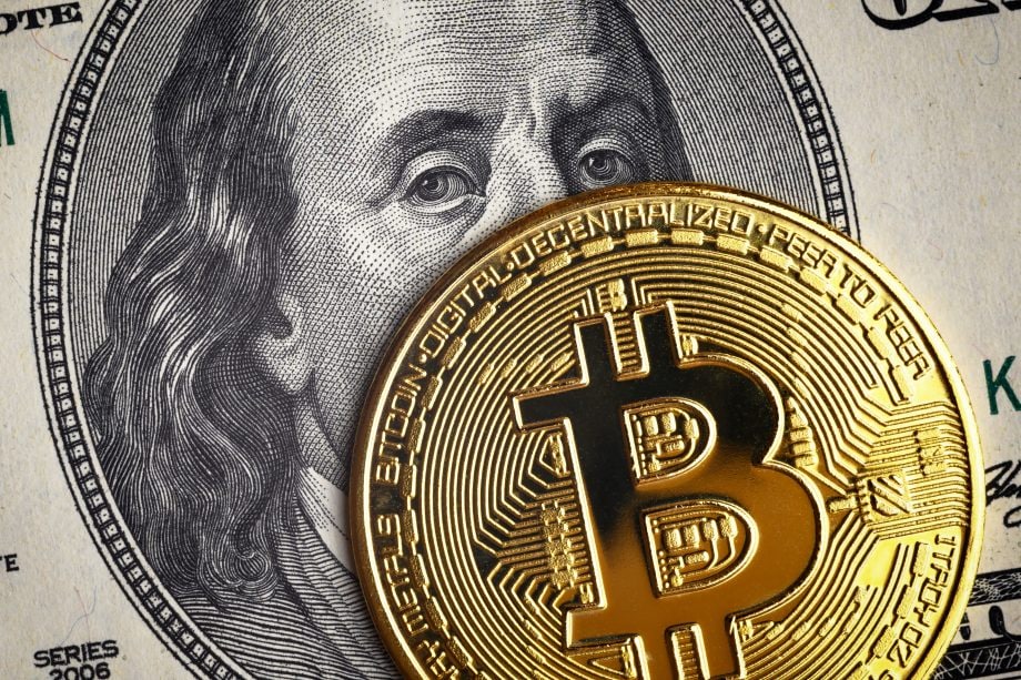 Eine goldene Bitcoin-Münze bedeckt teilweise einen US-Hundert-Dollar-Schein mit dem Gesicht Benjamin Franklins und symbolisiert damit die Überschneidung von Krypto und traditionellen Finanzen.