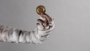 Das Liquiditätsgespenst macht Millionen mit Bitcoin Eine mit schmutzigen Bandagen umwickelte Hand hält eine Goldmünze mit dem Bitcoin-Symbol hoch und fängt den düsteren Reiz von Krypto vor einem schlichten grauen Hintergrund ein.