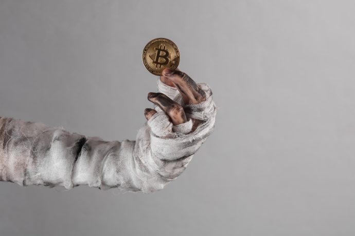 Eine mit schmutzigen Bandagen umwickelte Hand hält eine Goldmünze mit dem Bitcoin-Symbol hoch und fängt den düsteren Reiz von Krypto vor einem schlichten grauen Hintergrund ein.
