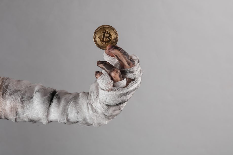 Eine mit schmutzigen Bandagen umwickelte Hand hält eine Goldmünze mit dem Bitcoin-Symbol hoch und fängt den düsteren Reiz von Krypto vor einem schlichten grauen Hintergrund ein.