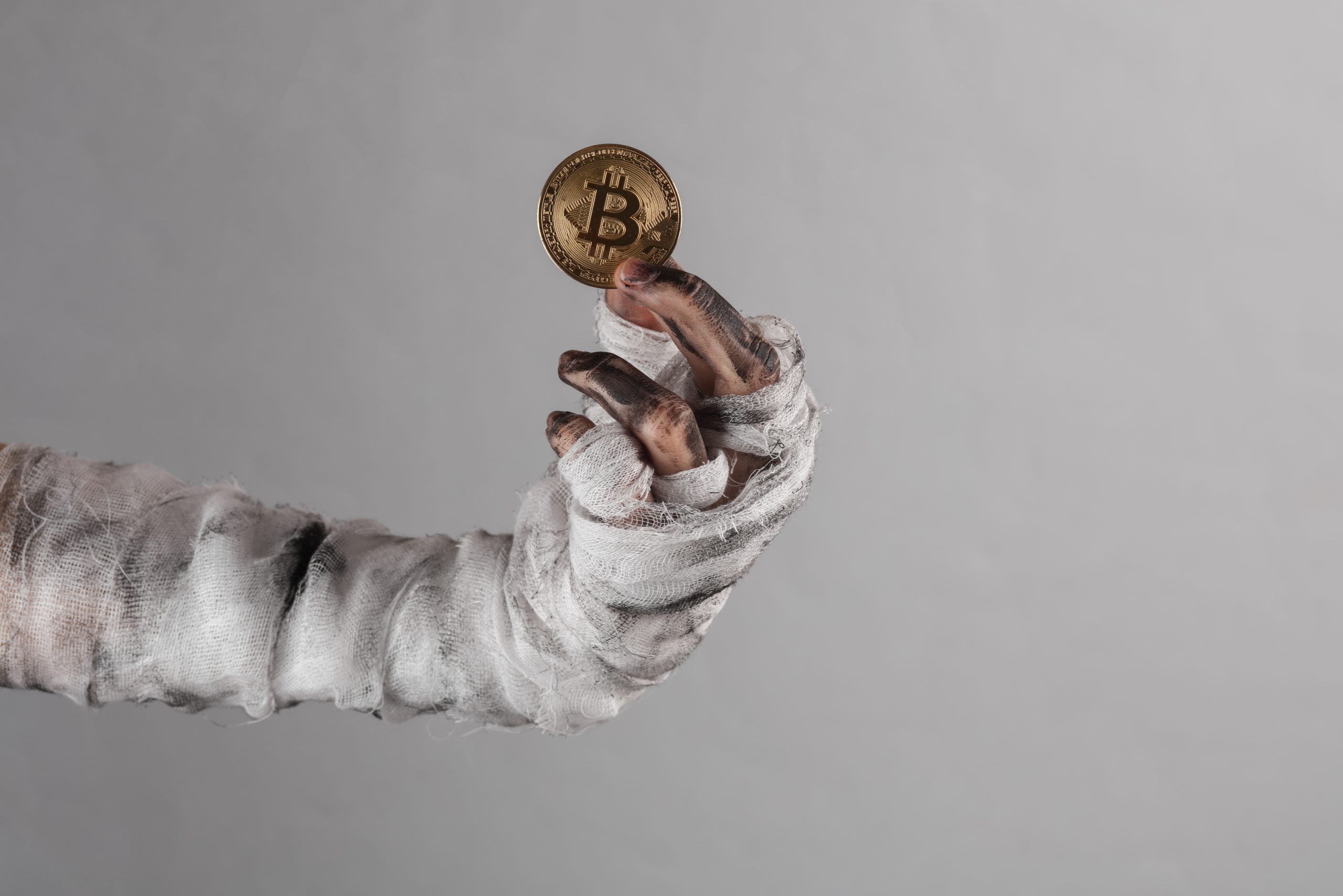 Eine mit schmutzigen Bandagen umwickelte Hand hält eine Goldmünze mit dem Bitcoin-Symbol hoch und fängt den düsteren Reiz von Krypto vor einem schlichten grauen Hintergrund ein.