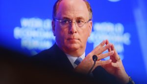 Larry Fink im Anzug und mit Brille sitzt mit verschränkten Fingern nachdenklich vor einem blauen Hintergrund mit verschwommenem weißen Text und BlackRock-Logos.