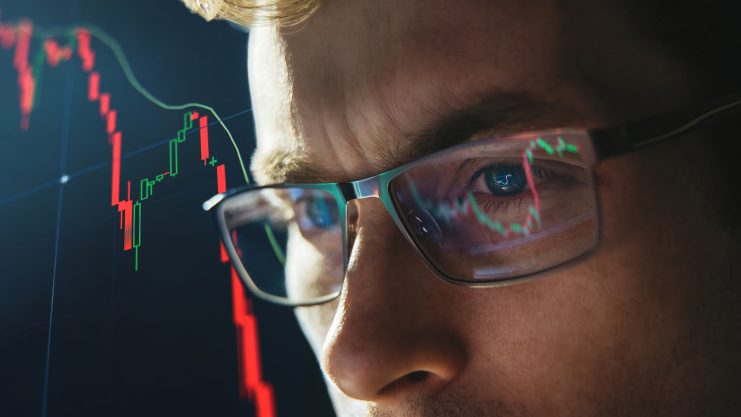 Nahaufnahme eines Händlers, der eine Brille trägt und ein Börsenchart mit rückläufigen roten Kerzenleuchtern analysiert, die sich in den Gläsern spiegeln.