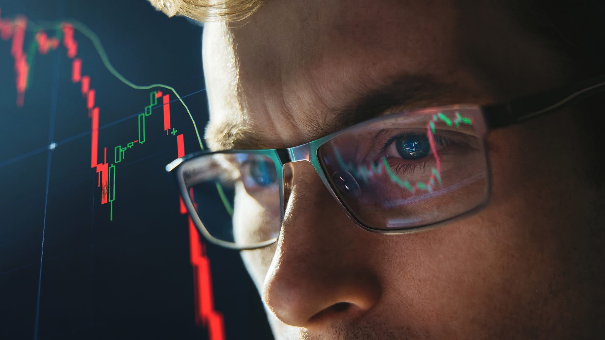 Nahaufnahme eines Händlers, der eine Brille trägt und ein Börsenchart mit rückläufigen roten Kerzenleuchtern analysiert, die sich in den Gläsern spiegeln.