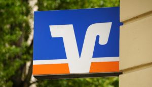 Ein blaues Schild mit einem weißen und orangefarbenen Logo mit stilisierten Buchstaben, das an der Seite eines beigen Gebäudes mit grünen Bäumen im Hintergrund angebracht ist.