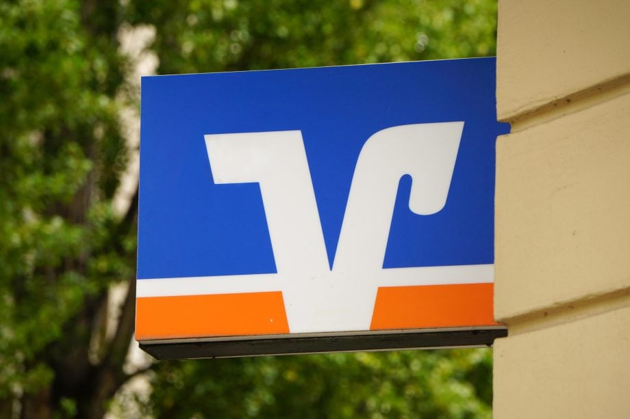 Ein blaues Schild mit einem weißen und orangefarbenen Logo mit stilisierten Buchstaben, das an der Seite eines beigen Gebäudes mit grünen Bäumen im Hintergrund angebracht ist.