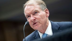 Ein Mann im Anzug spricht in ein Mikrofon bei einer scheinbar formellen Veranstaltung oder Anhörung, bei der es möglicherweise um Fragen im Zusammenhang mit Krypto geht.