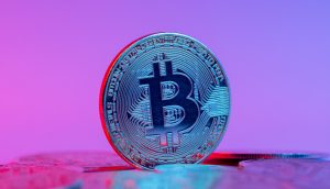 Eine physische Bitcoin-Münze steht aufrecht zwischen anderen Münzen vor einem Hintergrund mit rosa und lila Farbverlauf, der die einzigartige Präsenz von Bitcoin in der Welt der digitalen Währungen hervorhebt.