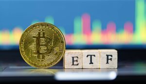 Eine physische Bitcoin-Münze steht neben Holzblöcken, die ETF buchstabieren, mit einer bunten Bitcoin-Finanztabelle im Hintergrund.