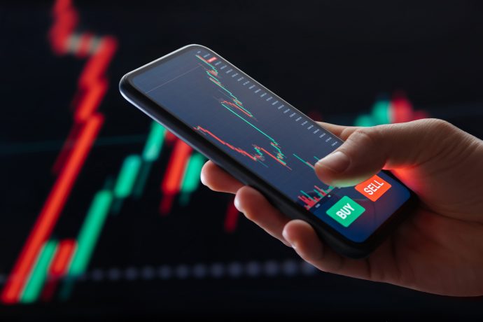 Eine Hand hält ein Smartphone, auf dem Krypto-Trades mit Hebel auf einer Aktienhandels-App angezeigt wird, mit einem roten und grünen Candlestick-Chart, Kauf- und Verkaufstasten, mit einem Finanzdiagramm im Hintergrund.