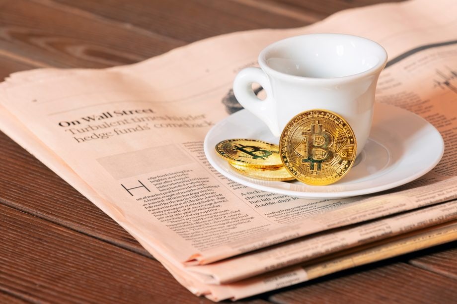 Eine weiße Kaffeetasse steht auf einer Untertasse auf einer Zeitung, daneben liegen zwei physische Bitcoin-Münzen und ein Hauch von Krypto-Intrige.