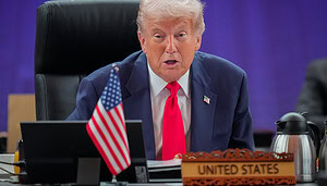 Donald Trump im Anzug und mit roter Krawatte sitzt an einem Konferenztisch mit einem Namensschild der Vereinigten Staaten und einer kleinen amerikanischen Flagge und diskutiert im Namen der US-Regierung über die Bitcoin-Politik, während er in ein Mikrofon spricht.