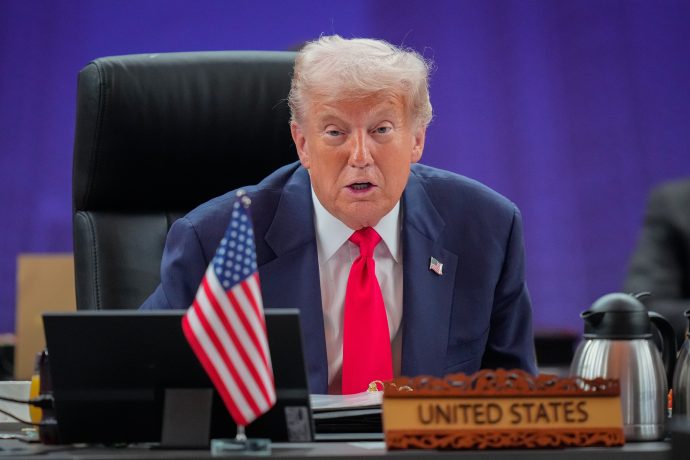 Trump Asia Donald Trump im Anzug und mit roter Krawatte sitzt an einem Konferenztisch mit einem Namensschild der Vereinigten Staaten und einer kleinen amerikanischen Flagge und diskutiert im Namen der US-Regierung über die Bitcoin-Politik, während er in ein Mikrofon spricht.