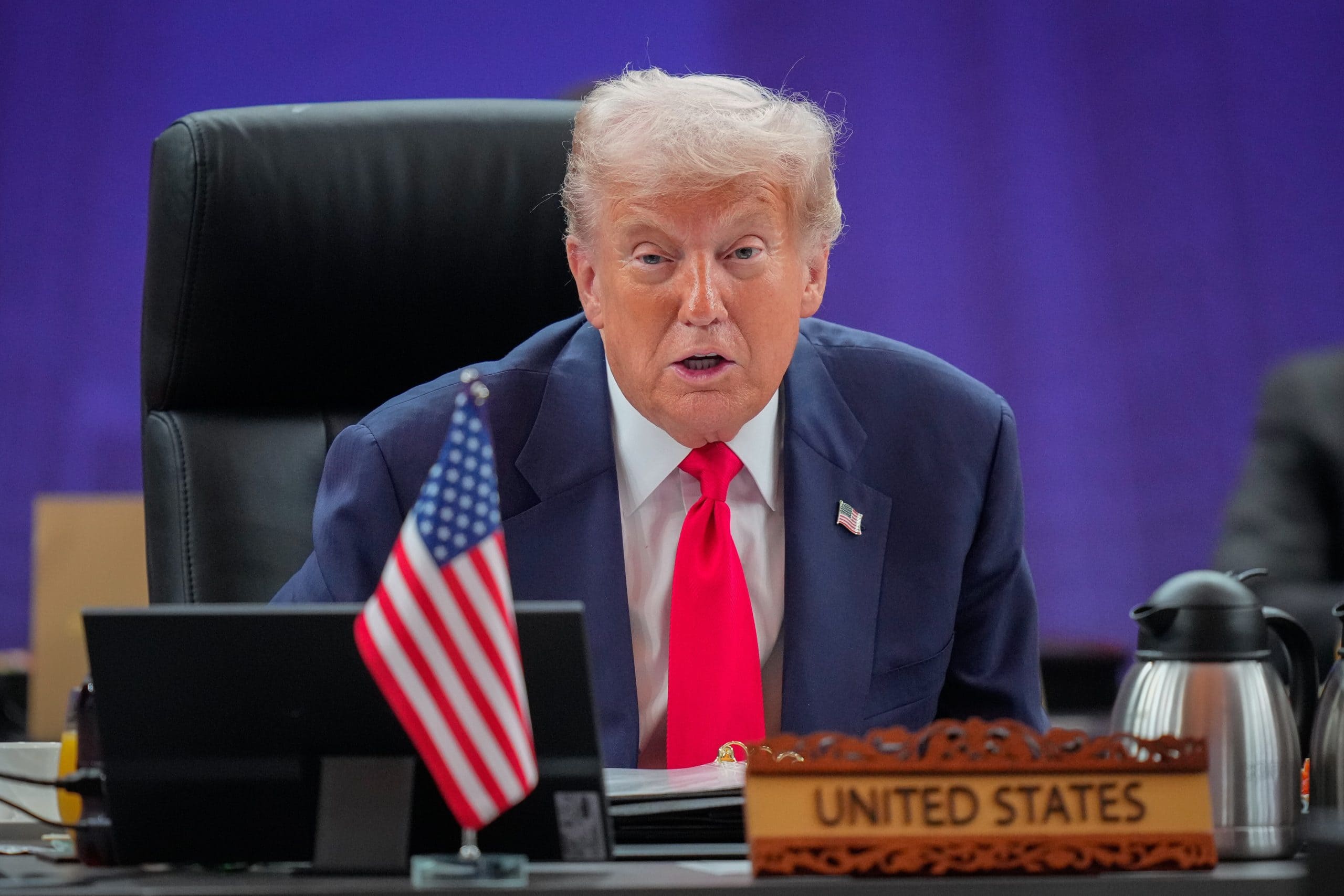 Trump Asia Donald Trump im Anzug und mit roter Krawatte sitzt an einem Konferenztisch mit einem Namensschild der Vereinigten Staaten und einer kleinen amerikanischen Flagge und diskutiert im Namen der US-Regierung über die Bitcoin-Politik, während er in ein Mikrofon spricht.