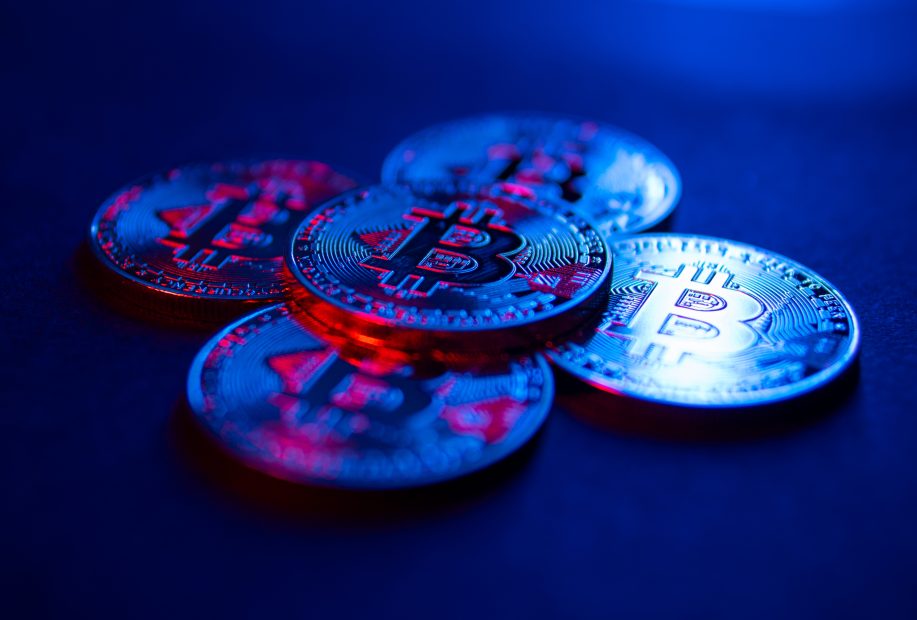 Fünf metallische Bitcoin-Münzen sind auf einer dunklen Fläche angeordnet, die mit blauem und rotem Licht beleuchtet wird und an die High-Tech-Welt der Kryptoanalyse und Unternehmen wie Chainalysis erinnert.