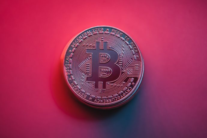 Eine metallische Bitcoin-Münze ruht auf einem Hintergrund mit rosa und violettem Farbverlauf, der das ikonische Bitcoin-Symbol und verschlungene kreislaufartige Muster zeigt.