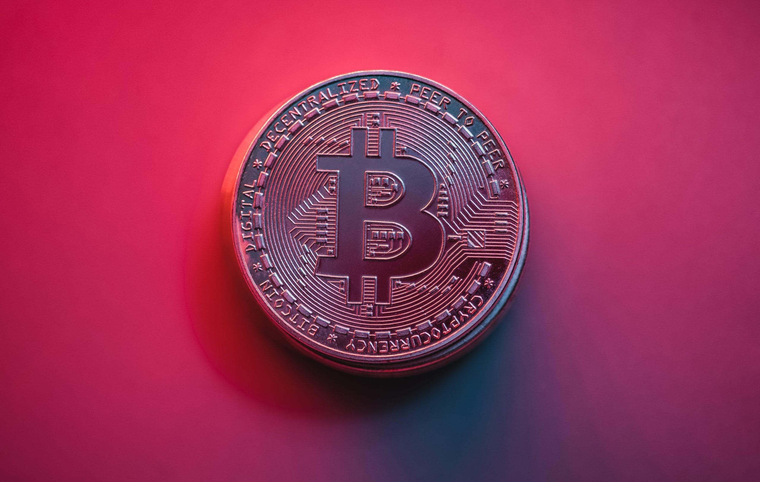 Eine metallische Bitcoin-Münze ruht auf einem Hintergrund mit rosa und violettem Farbverlauf, der das ikonische Bitcoin-Symbol und verschlungene kreislaufartige Muster zeigt.