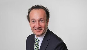 Dr.-Robert-Bosch-scaled Ein Mann in einem dunklen Anzug, weißem Hemd und grün gestreifter Krawatte lächelt vor einem schlichten hellen Hintergrund in die Kamera und trägt dezent eine goldene Bitcoin-Anstecknadel.