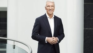 Ralf Wintergerst in dunklem Anzug und weißem Hemd steht drinnen an einem Metallgeländer und lächelt vor einem weiß-schwarzen Hintergrund in die Kamera - möglicherweise auf einer Veranstaltung von Giesecke+Devrient zum Digitalen Euro.