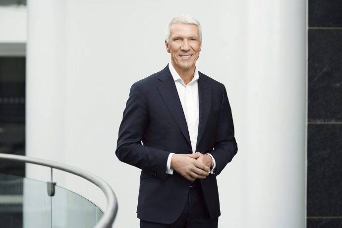 Ralf Wintergerst in dunklem Anzug und weißem Hemd steht drinnen an einem Metallgeländer und lächelt vor einem weiß-schwarzen Hintergrund in die Kamera - möglicherweise auf einer Veranstaltung von Giesecke+Devrient zum Digitalen Euro.