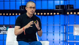 Changpeng Zhao in einem schwarzen Polohemd und einer blauen Hose spricht auf einem weißen Stuhl sitzend vor einem blau beleuchteten Bühnenhintergrund über Binance und die jüngste französische Behördenkontrolle.