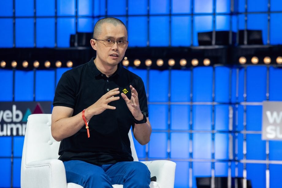 Changpeng Zhao in einem schwarzen Polohemd und einer blauen Hose spricht auf einem weißen Stuhl sitzend vor einem blau beleuchteten Bühnenhintergrund über Binance und die jüngste französische Behördenkontrolle.