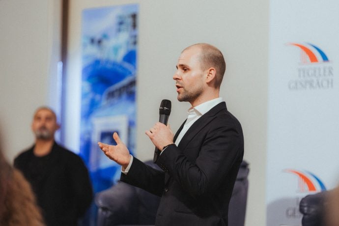 Ein Mann im Anzug spricht während einer Indoor-Veranstaltung in ein Handmikrofon über Bitcoin, wobei ein Teilnehmer und Banner im Hintergrund verschwimmen.