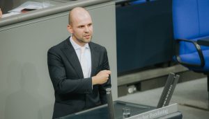 Ein Mann im Anzug steht an einem Podium mit der Aufschrift Deutscher Bundestag und spricht über Bitcoin oder gestikuliert mit seinem rechten Arm in einer formellen Umgebung.