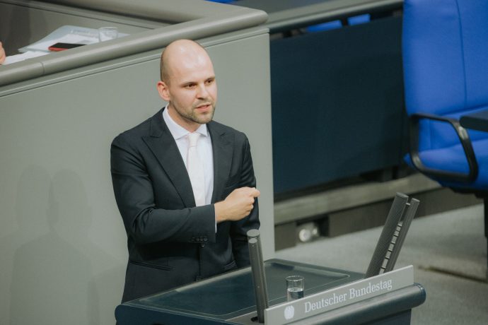 Ein Mann im Anzug steht an einem Podium mit der Aufschrift Deutscher Bundestag und spricht über Bitcoin oder gestikuliert mit seinem rechten Arm in einer formellen Umgebung.