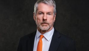 Ein Mann mittleren Alters mit grauem Haar und Bart, der einen schwarzen Anzug, ein weißes Hemd und eine orangefarbene Krawatte trägt, steht vor einem dunklen Hintergrund.