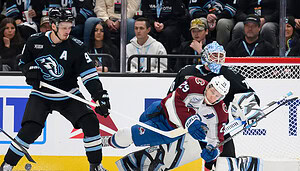 Ein Eishockeyspieler in einem weißen Trikot versucht, ein Tor zu erzielen, während ein Torwart und ein Verteidiger in schwarzen Trikots das Netz während eines Spiels blockieren, während im Hintergrund Zuschauer und Polymarket-Fans zusehen.