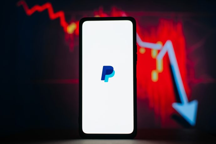 Auf einem Smartphone wird das PayPal-Logo angezeigt, mit einem roten, abwärts gerichteten Aktienchart und einem blauen Pfeil im Hintergrund.