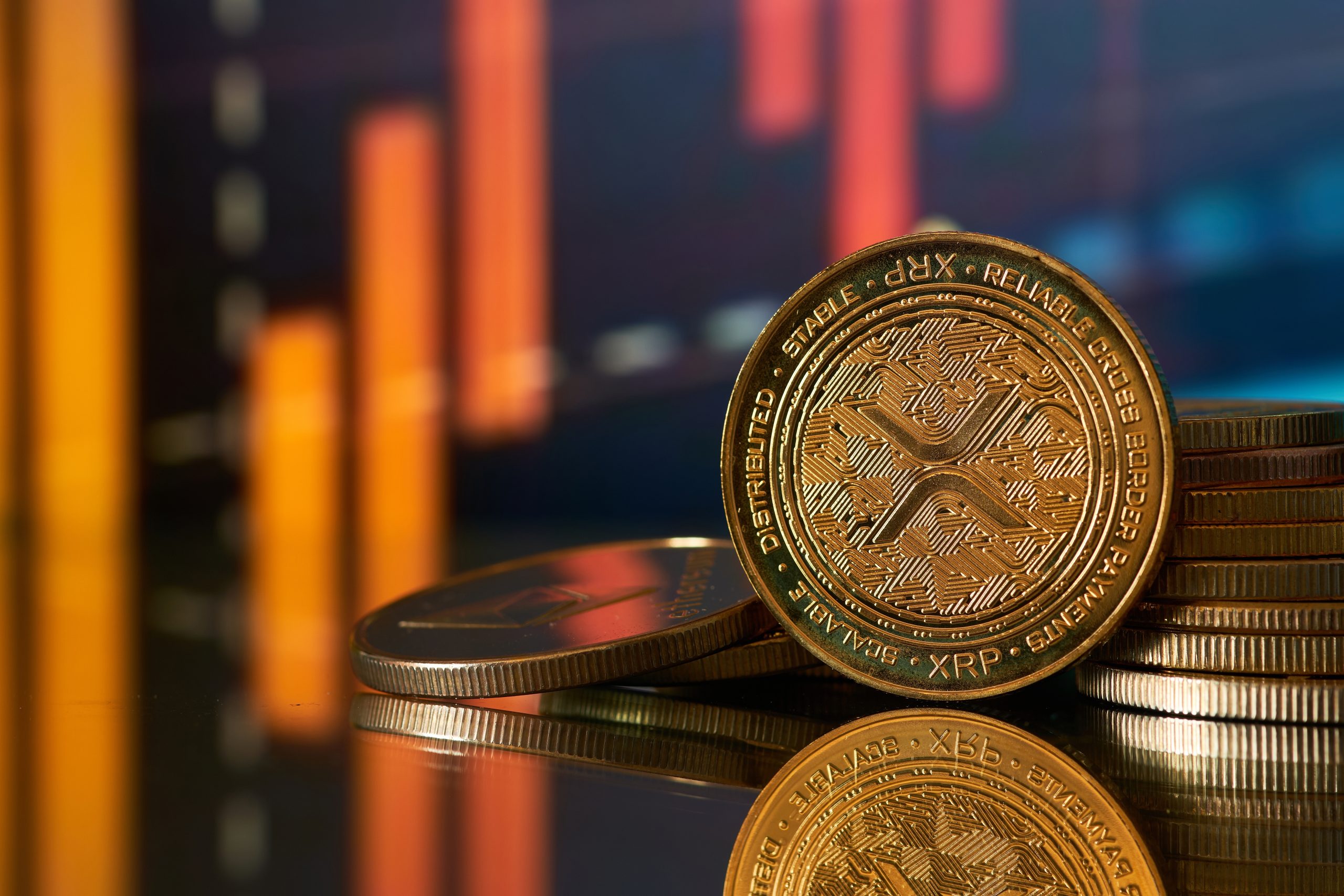 Nahaufnahme von gestapelten XRP-Kryptowährungsmünzen mit orangefarbenem Balkendiagramm im unscharfen Hintergrund, was auf Finanzdaten oder Markttrends hindeutet.
