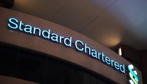 Das beleuchtete Logo und der Name von Standard Chartered sind nachts auf der Außenseite eines Gebäudes zu sehen und unterstreichen die prominente Partnerschaft mit globalen Unternehmen.