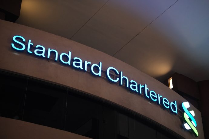 Das beleuchtete Logo und der Name von Standard Chartered sind nachts auf der Außenseite eines Gebäudes zu sehen und unterstreichen die prominente Partnerschaft mit globalen Unternehmen.